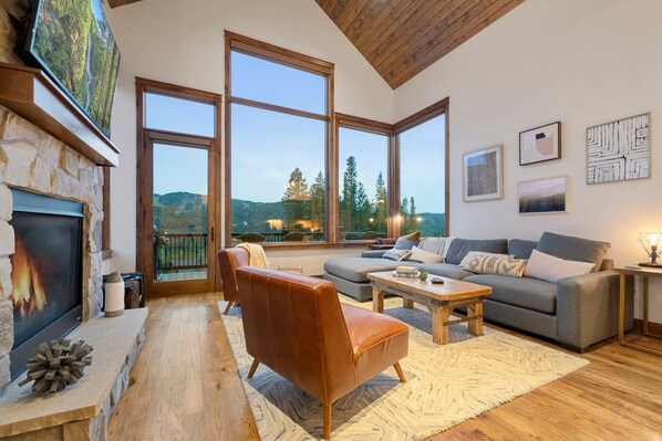 Amplia sala de estar con chimenea de piedra, ventanas grandes que ofrecen vistas al exterior, un sofá confortable y una mesa de centro.