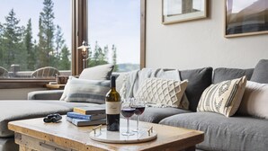 Ein gemütliches Wohnzimmer mit einem Holztisch, einer Weinflasche und Gläsern darauf. Ein graues Sofa mit Kissen und ein Fenster mit Blick auf Bäume.