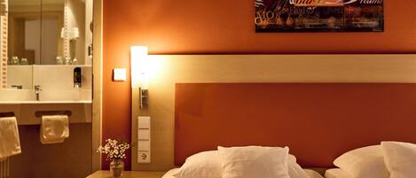 A hotel room with a bed, a nightstand with a lamp, a vase with flowers, and a wall-mounted artwork.