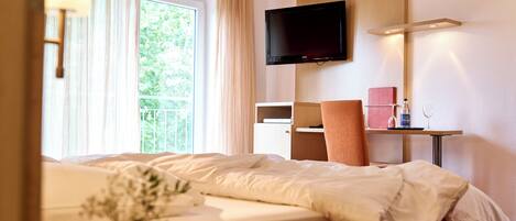 A hotel room with a bed, a TV mounted on the wall, a small table with a bottle and glass, and a window with curtains.