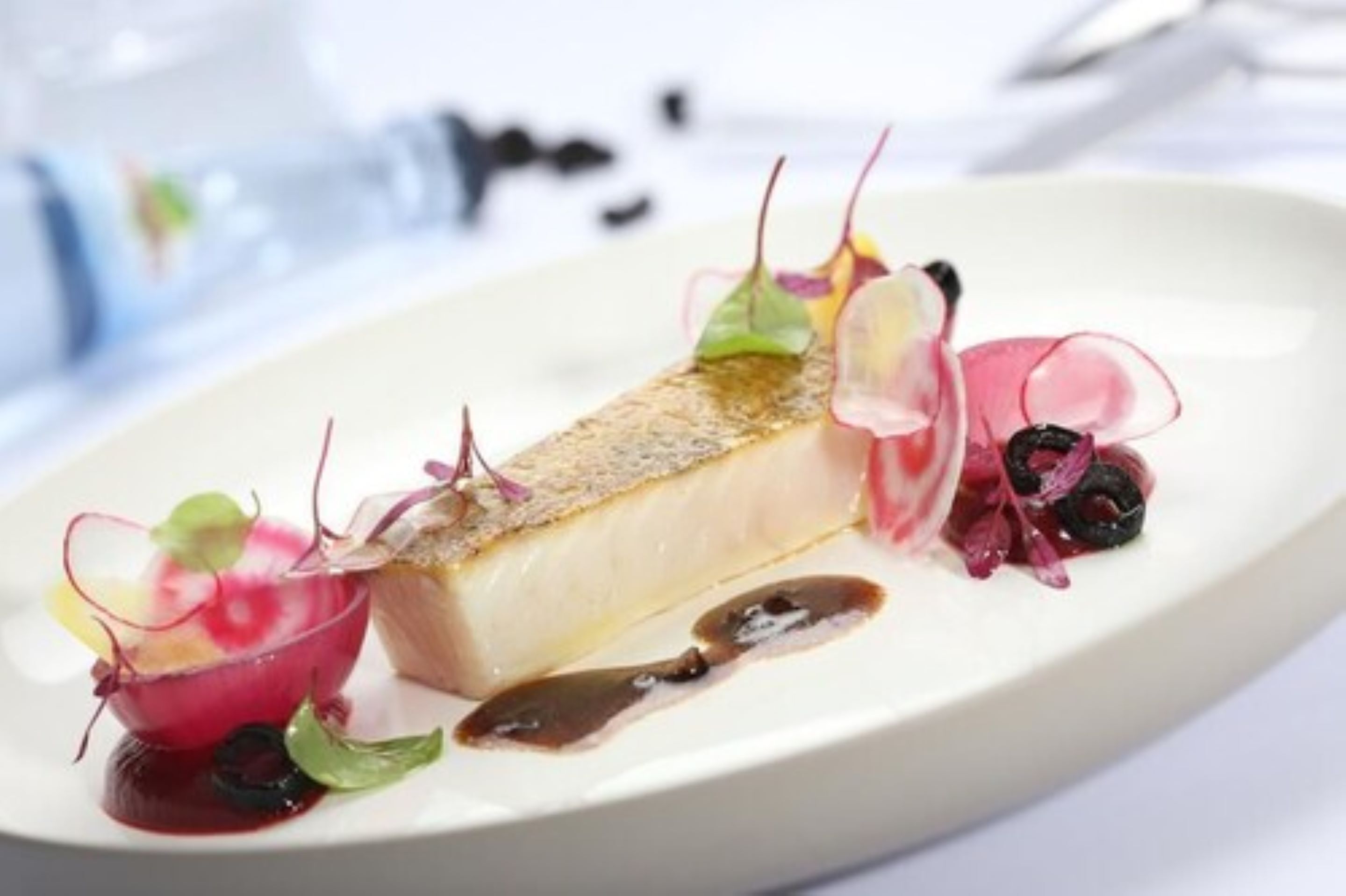 A white plate with a piece of fish, garnished with red and green vegetables and a dark sauce.