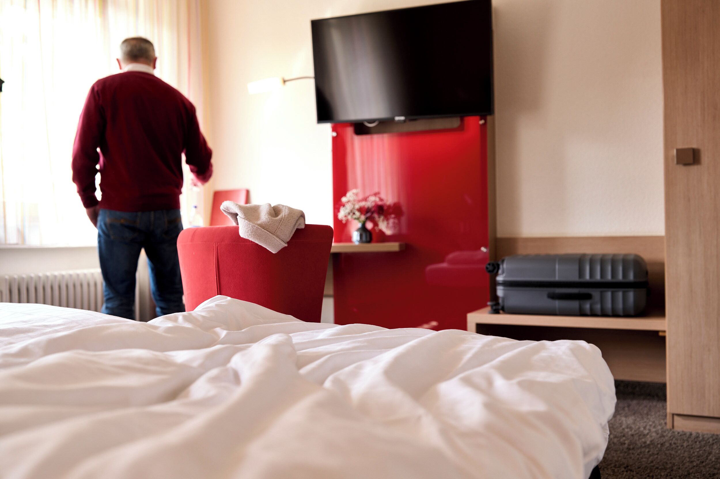 Un cuarto de hotel con una cama, un sillón rojo, un televisor de pantalla plana montado en la pared, una mesita de noche de madera y una maleta en el suelo.