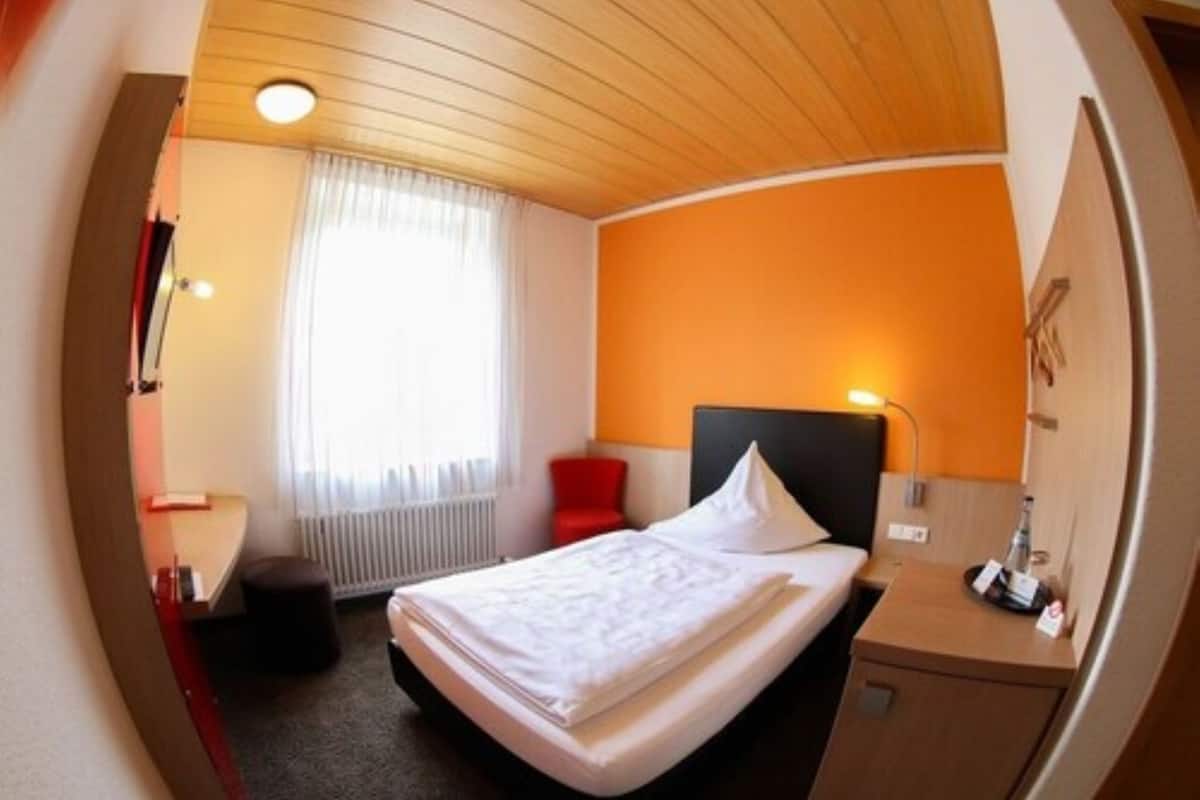 A hotel room with a bed, a bedside table, a red chair, a window with curtains, and a wooden ceiling.