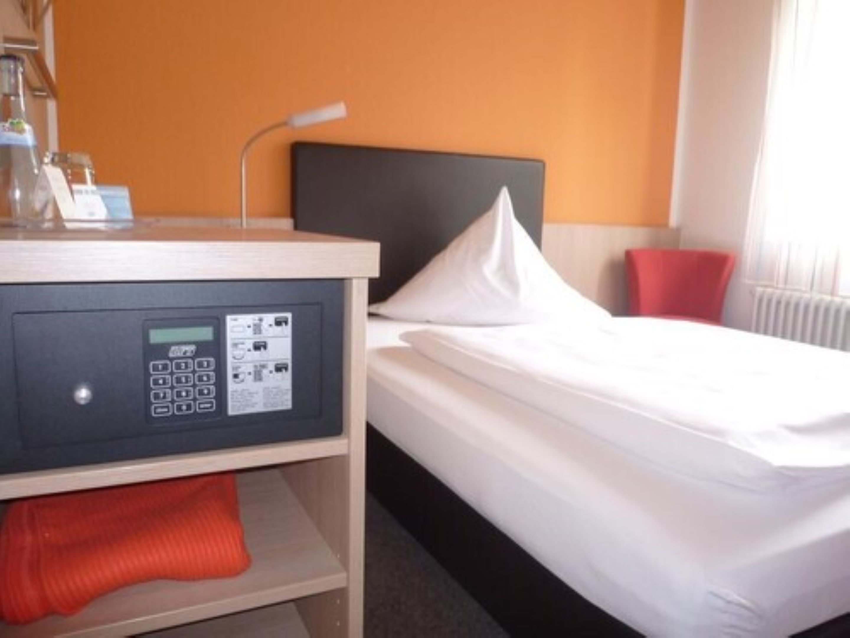 Una habitación de hotel con cama, panel de control, almohada roja y ventana con persianas.