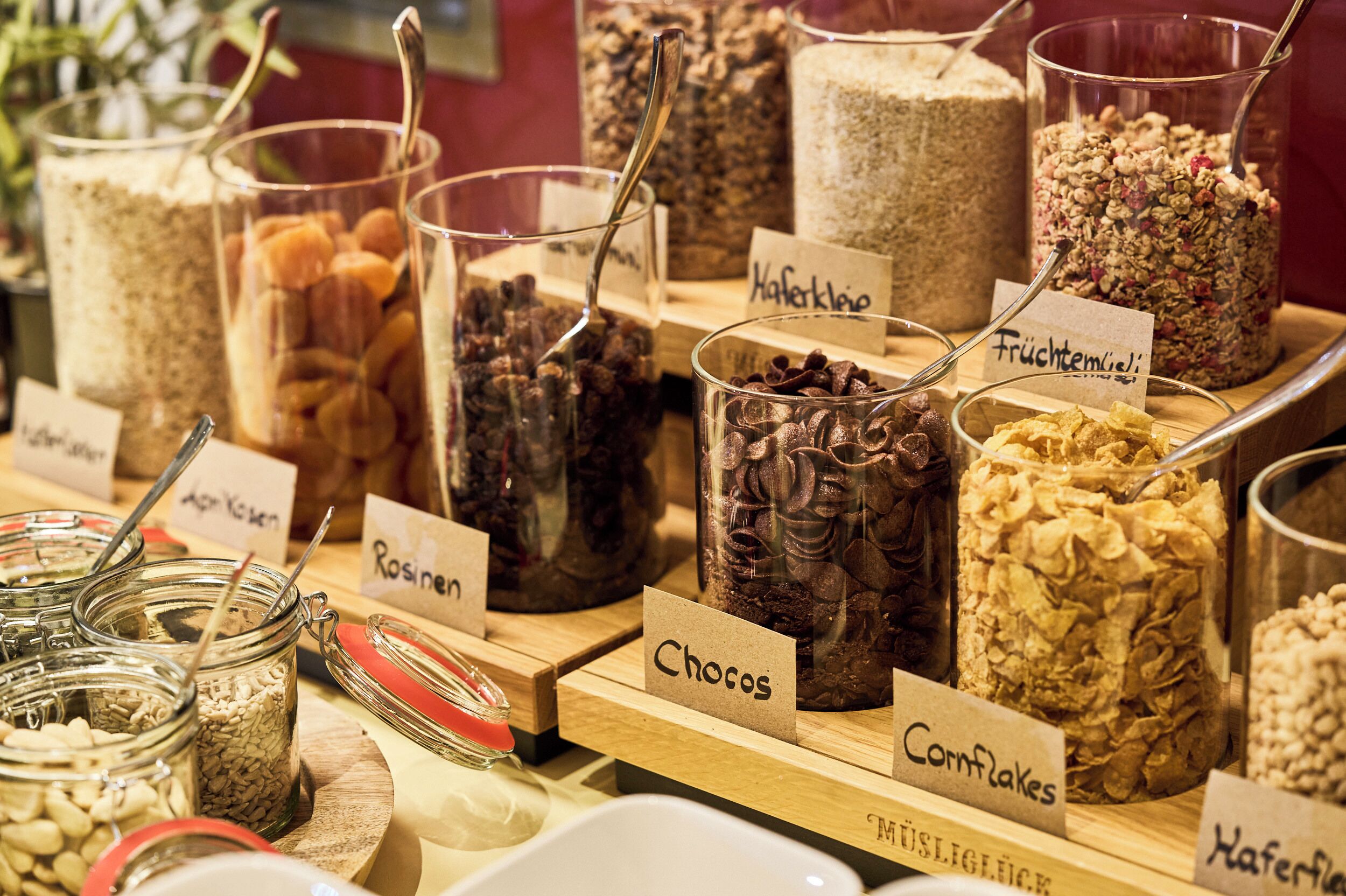 A variety of food items in glass containers with labels, including nuts, chocolate, and cornflakes.
