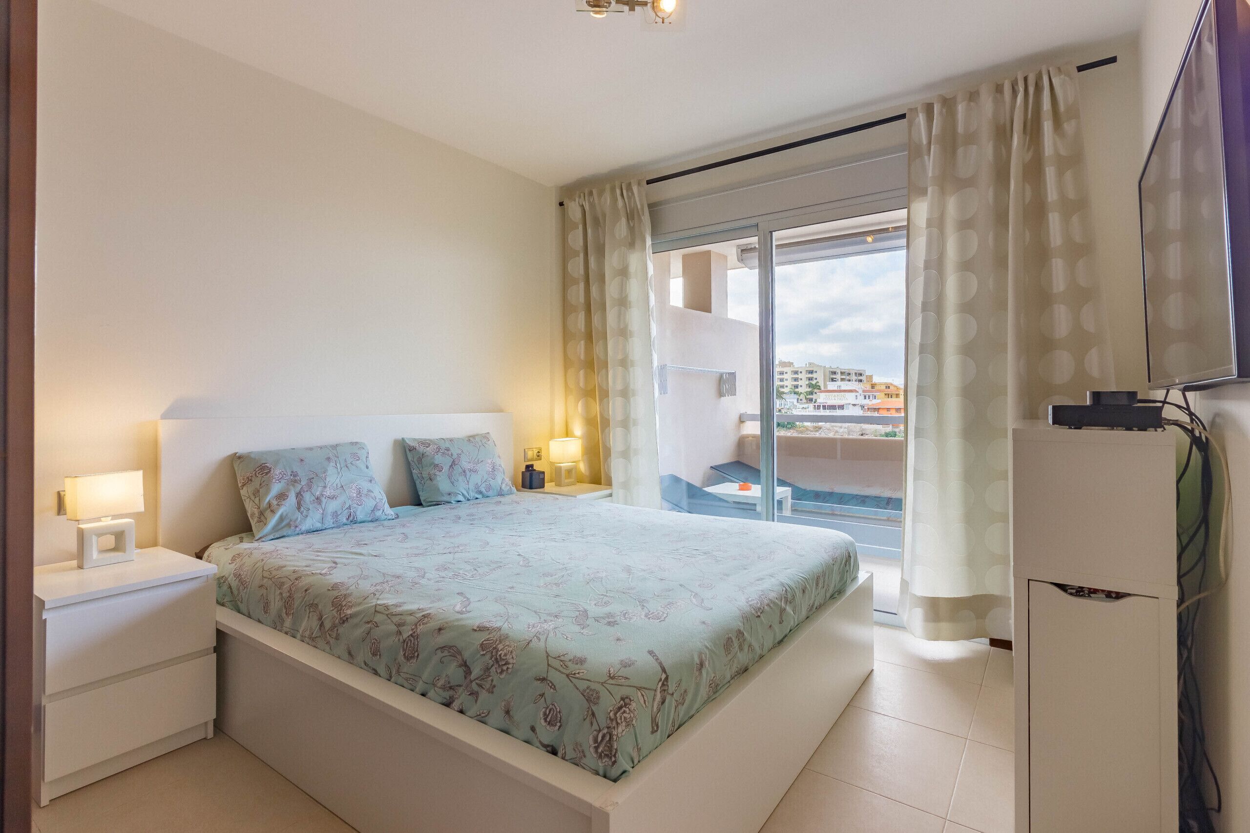 A bedroom with a bed, bedside lamps, a TV, and a view of the city through a large window.