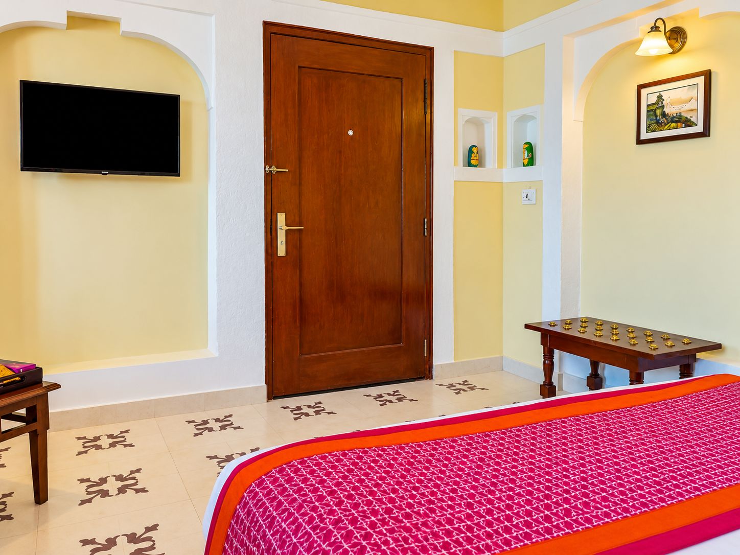 Una habitación de hotel con una puerta de madera, un televisor de pantalla plana montado en la pared, una mesita con objetos decorativos y una cama con edredón estampado.