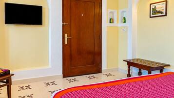 Una habitación de hotel con una puerta de madera, un televisor de pantalla plana montado en la pared, una mesita con objetos decorativos y una cama con edredón estampado.