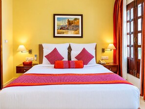 A hotel room with a bed, two bedside tables, a nightstand, a wall-mounted lamp, a framed picture, and a window with curtains. - Guleria Kothi (Varanasi)