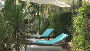 A sunlit outdoor lounge area with two wooden lounge chairs, one with a white cushion and the other with a blue cushion, under a large umbrella.