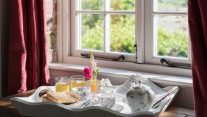 A breakfast setup on a tray by a window with a view of greenery, including a vase with flowers, a teapot, cups, and a glass of orange juice. - Award winning stunning barn conversion over looking nature reserve (Landulph)