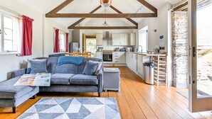 A modern living room with a grey sofa, wooden beams, a kitchen in the background, and a stone wall. - Award winning stunning barn conversion over looking nature reserve (Landulph)
