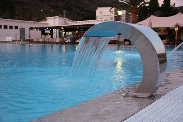 A swimming pool with a water feature, a lounge area with chairs, and a building in the background.