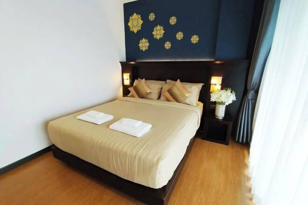 Een net opgemaakt bed met kussens en een gevouwen handdoek, een wand met decoratieve elementen en een nachtkastje met een vaas bloemen.