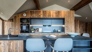 A modern kitchen with wooden accents, a built-in oven, a coffee maker, and a large island with bar stools. - MOULIN III - Luxury chalet with private hot tub (Les Gets)