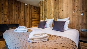 A hotel room with a wooden wall panel, a bed with white linens and dark pillows, a desk with a chair, and a small table with a lamp. - MOULIN III - Luxury chalet with private hot tub (Les Gets)