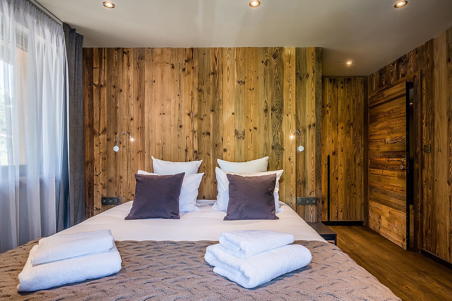 A modern bedroom with a wooden wall panel design, a large bed with white bedding and two pillows, and a pair of towels on the bed.