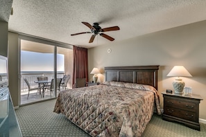 A hotel room with a bed, two bedside tables with lamps, a ceiling fan, a balcony with a table and chairs, and a view of the ocean. - Appealing Earth Toned Unit, 9th Floor, Hot Tub | Crescent Shores - S 908 (North Myrtle Beach)
