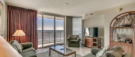 A spacious living room with a large sliding glass door leading to a balcony, a comfortable sofa set, a coffee table, and a television on a wooden stand.