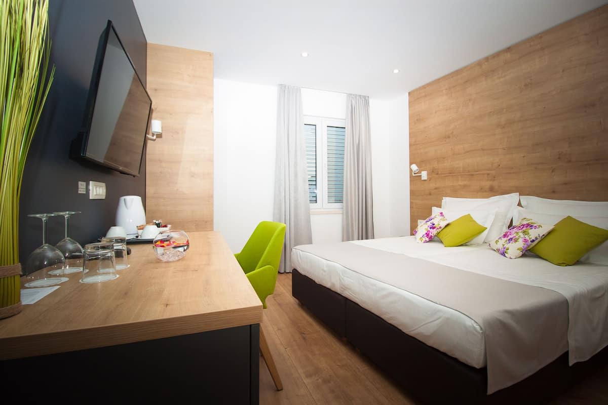 A modern hotel room with a wooden desk, a bed with multiple pillows, a green chair, and a TV mounted on the wall.