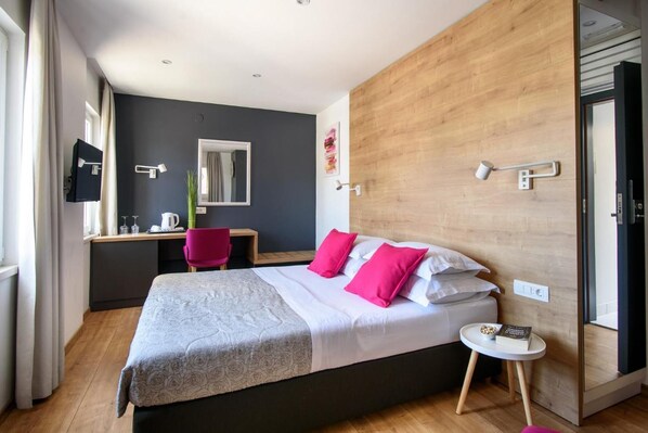 Quarto moderno com uma cama grande, uma escrivaninha e uma cadeira. Há um espelho na parede, uma janela com cortinas e uma mesinha de cabeceira com um livro.