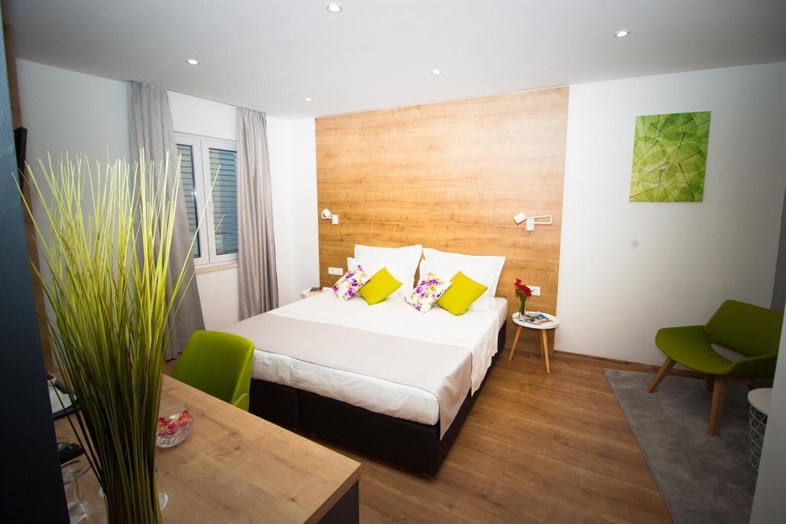 A modern hotel room with a wooden headboard, a large bed with white bedding and colorful pillows, a small round table with a vase of flowers, two green chairs, and a large green abstract painting on the wall.