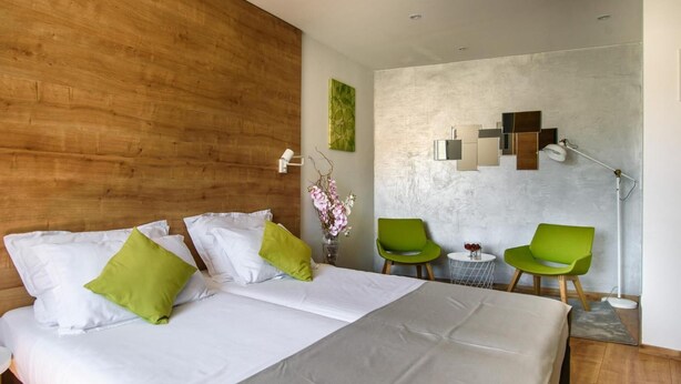 A hotel room with a wooden headboard, a bed with white and green pillows, a green chair, a small table with a vase of flowers, and a wall-mounted light fixture. - ORA Luxury Rooms (Dubrovnik)