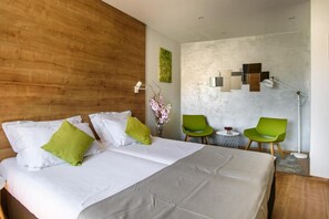 Quarto de hotel com cabeceira de madeira, cama com almofadas brancas e verdes, uma cadeira verde, uma mesa pequena com um vaso de flores e um aplique na parede.