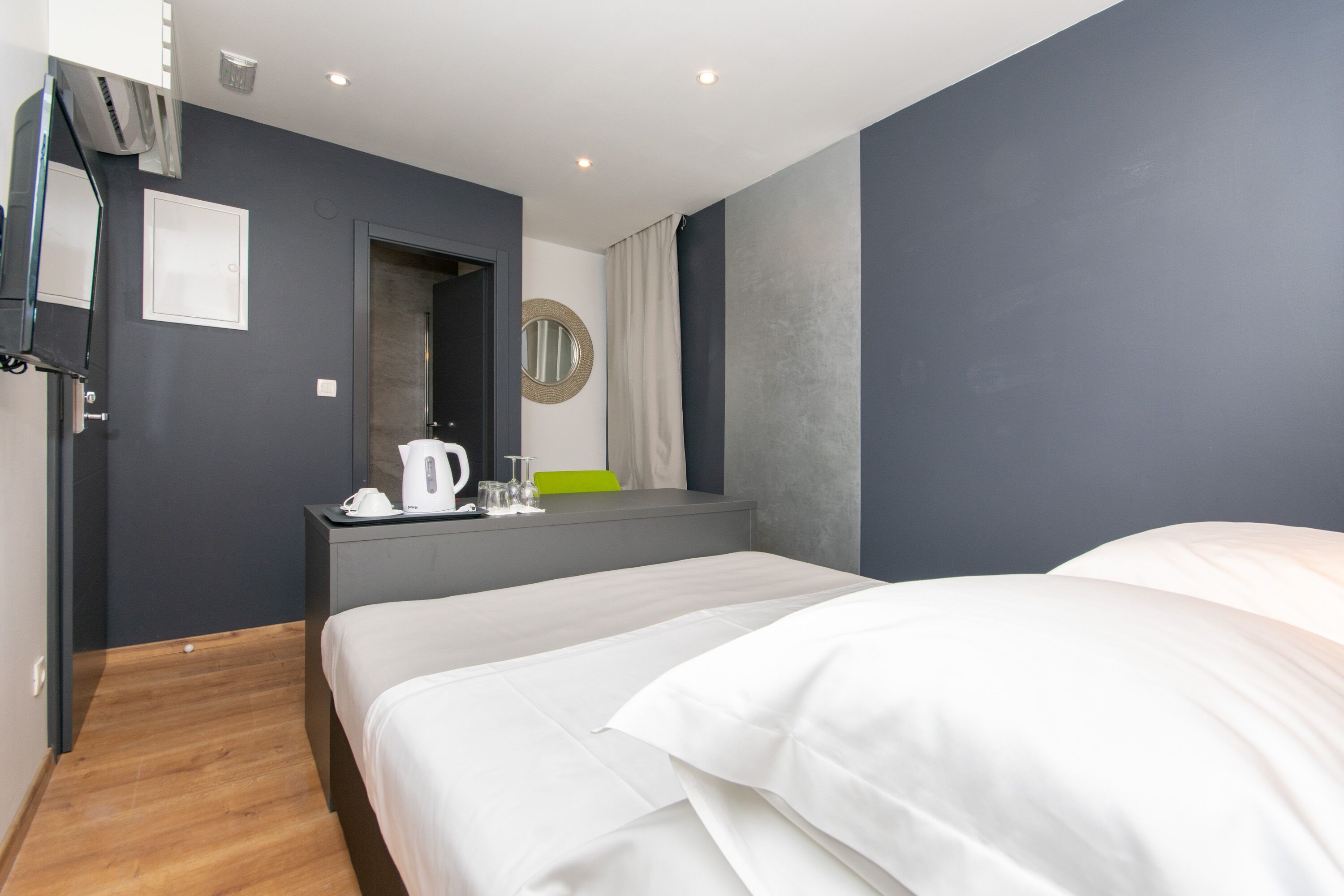 A modern hotel room with a large bed, a bedside table with a kettle, a mirror, and a view of the city.