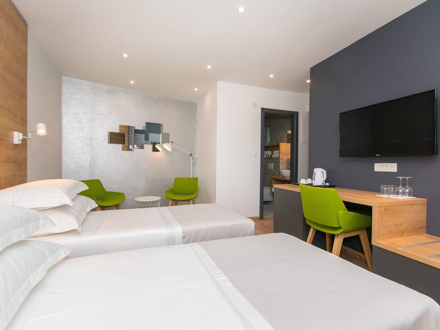Una habitación de hotel moderna con dos camas, un escritorio, un televisor y una zona de estar con sillas verdes.