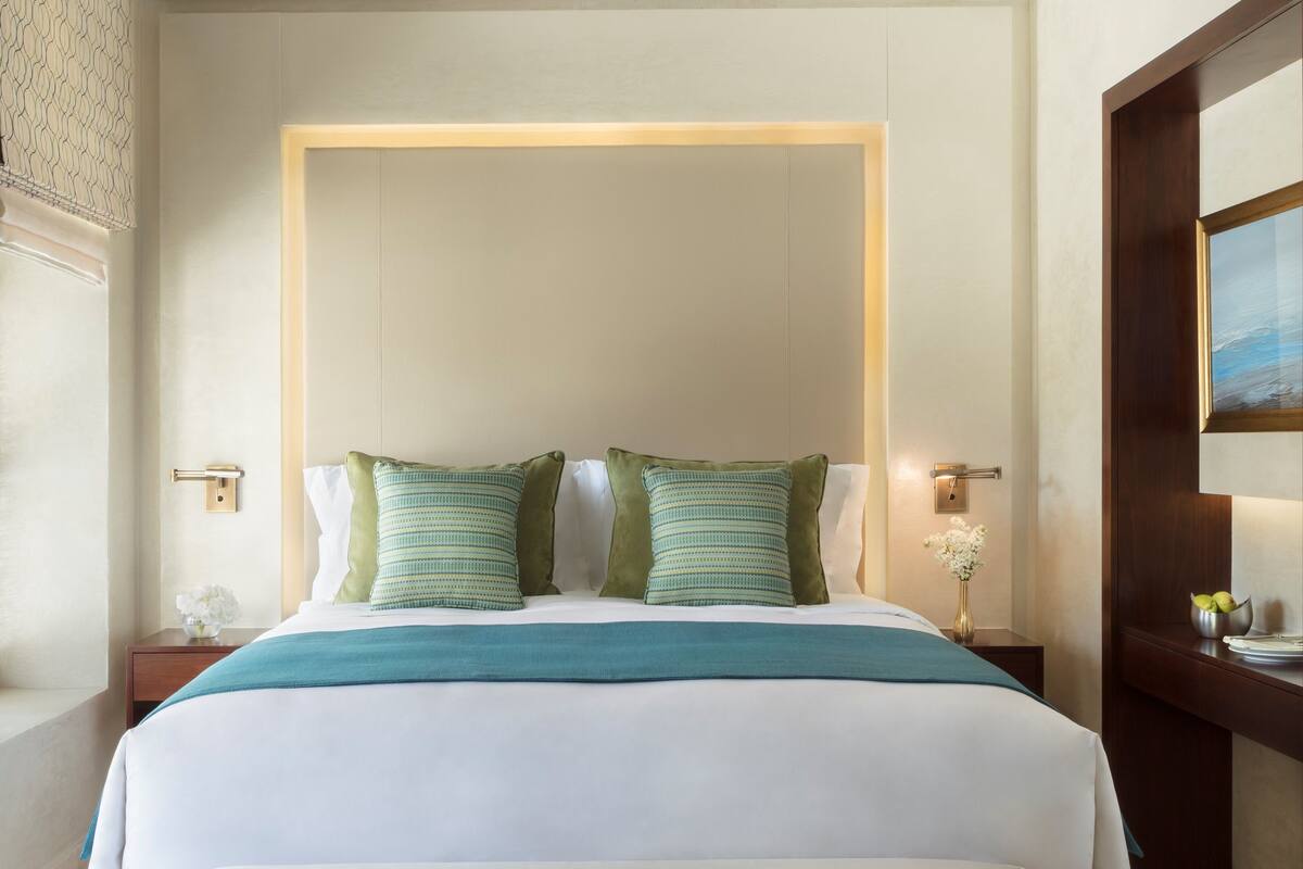 A hotel room with a large bed, two pillows, a nightstand with a vase, and a painting on the wall.