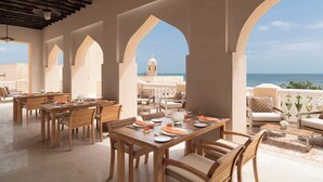 A covered outdoor dining area with wooden tables and chairs, arches, and a view of the sea. - Souq Al Wakra Hotel Qatar by Tivoli (Al Wakrah)