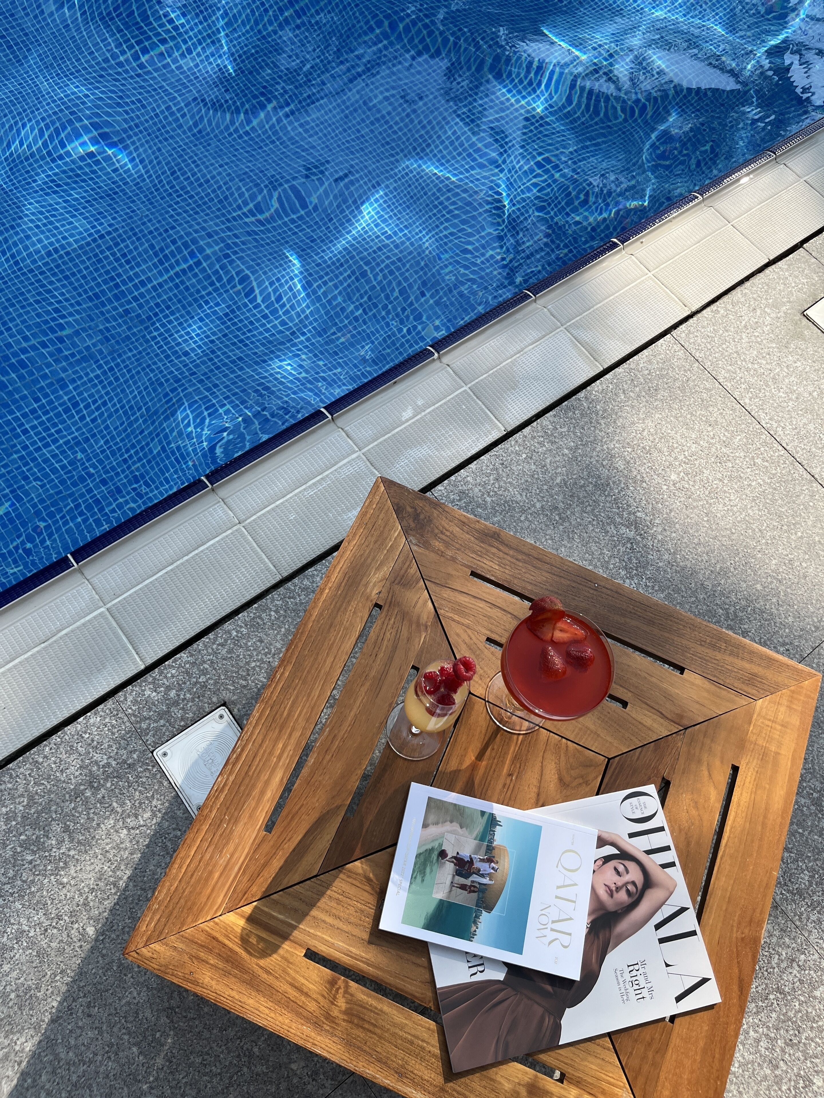 Una mesa de madera con una revista, una bebida con fresas y una fotografía cerca de una piscina.