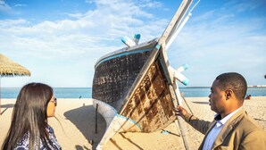 A beach scene with a wooden structure, possibly a boat, and a thatched umbrella. - Souq Al Wakra Hotel Qatar by Tivoli (Al Wakrah)