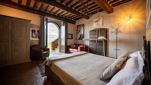 A bedroom with a large bed, a red sofa, a wooden wardrobe, and a view through a glass door. - Il Burellino Wine & Resort (Montalcino)