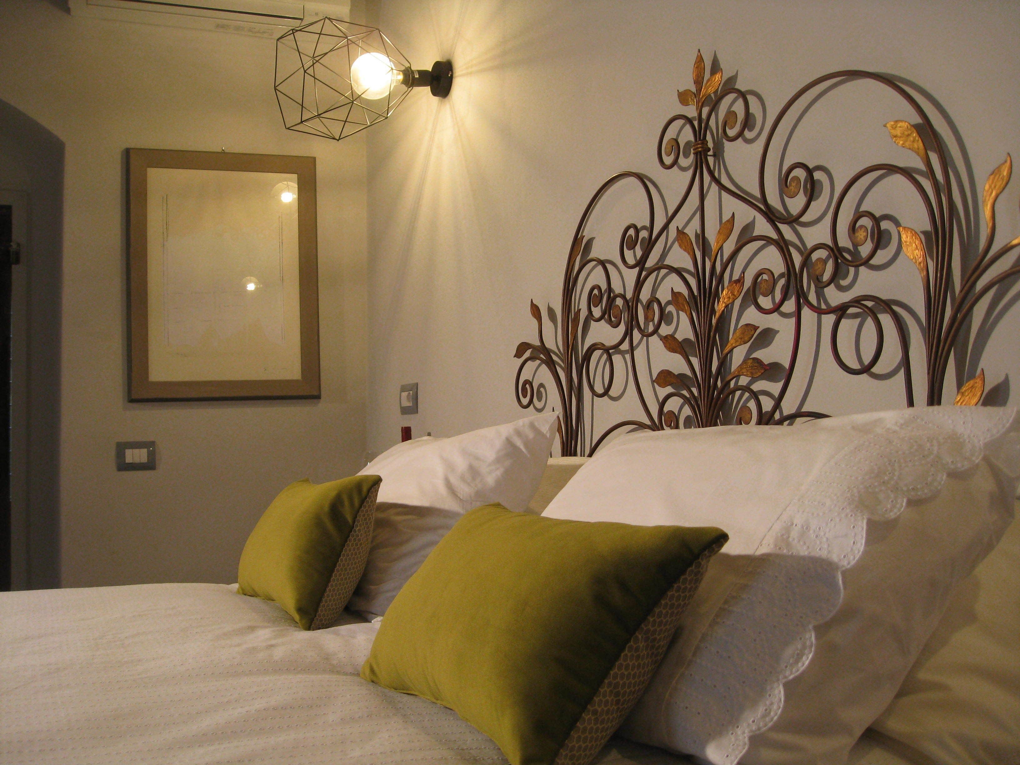 A neatly made bed with white linens and two green pillows, a wall-mounted light fixture, and a decorative metal wall art piece.