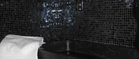 Un baño con pared de azulejos negros, un espejo, un lavabo y una vela.