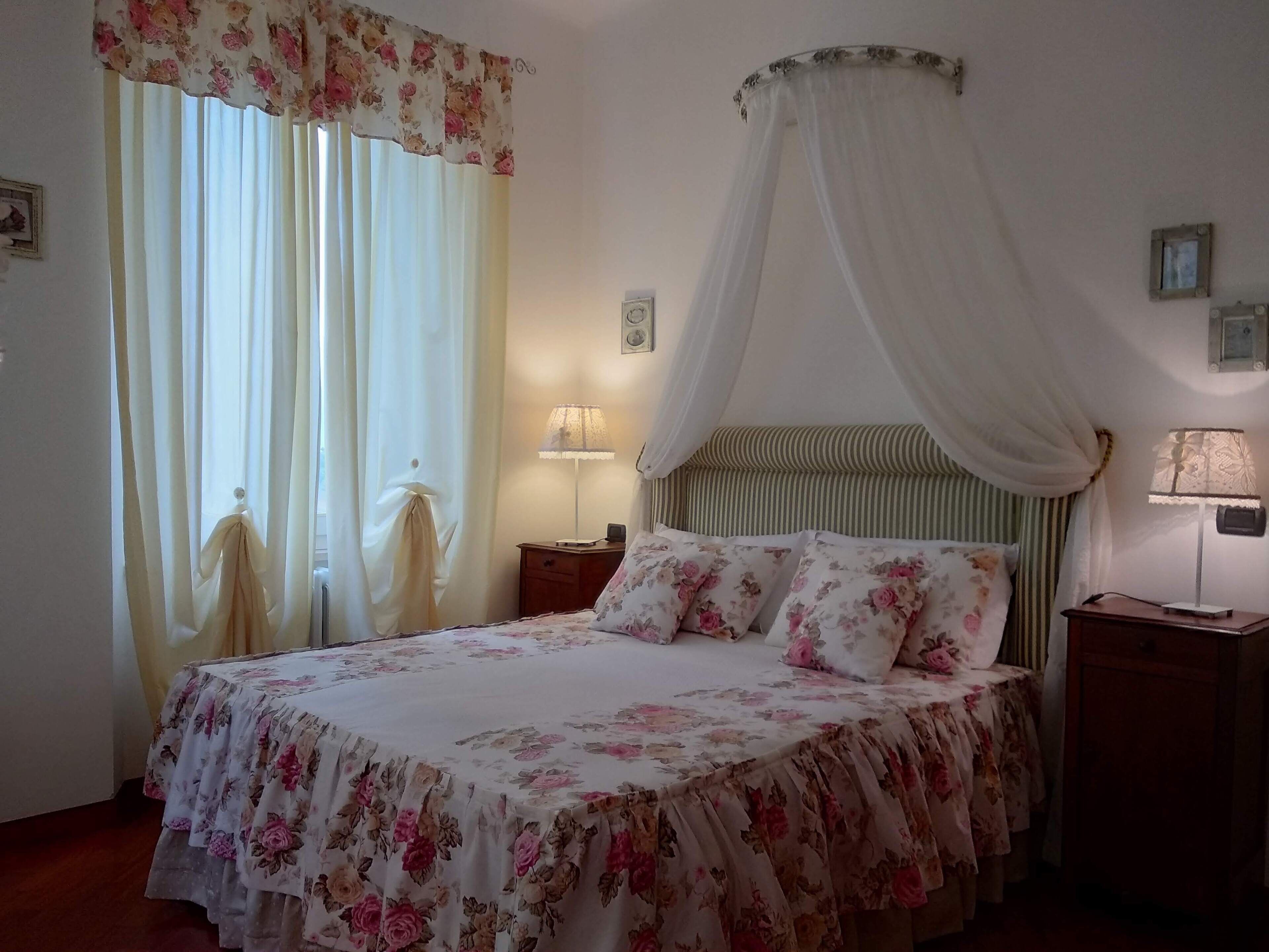 Ein Schlafzimmer mit einem Bett, blumigem Bettzeug, einem Baldachin, einem Nachttisch mit Lampe und Vorhängen.