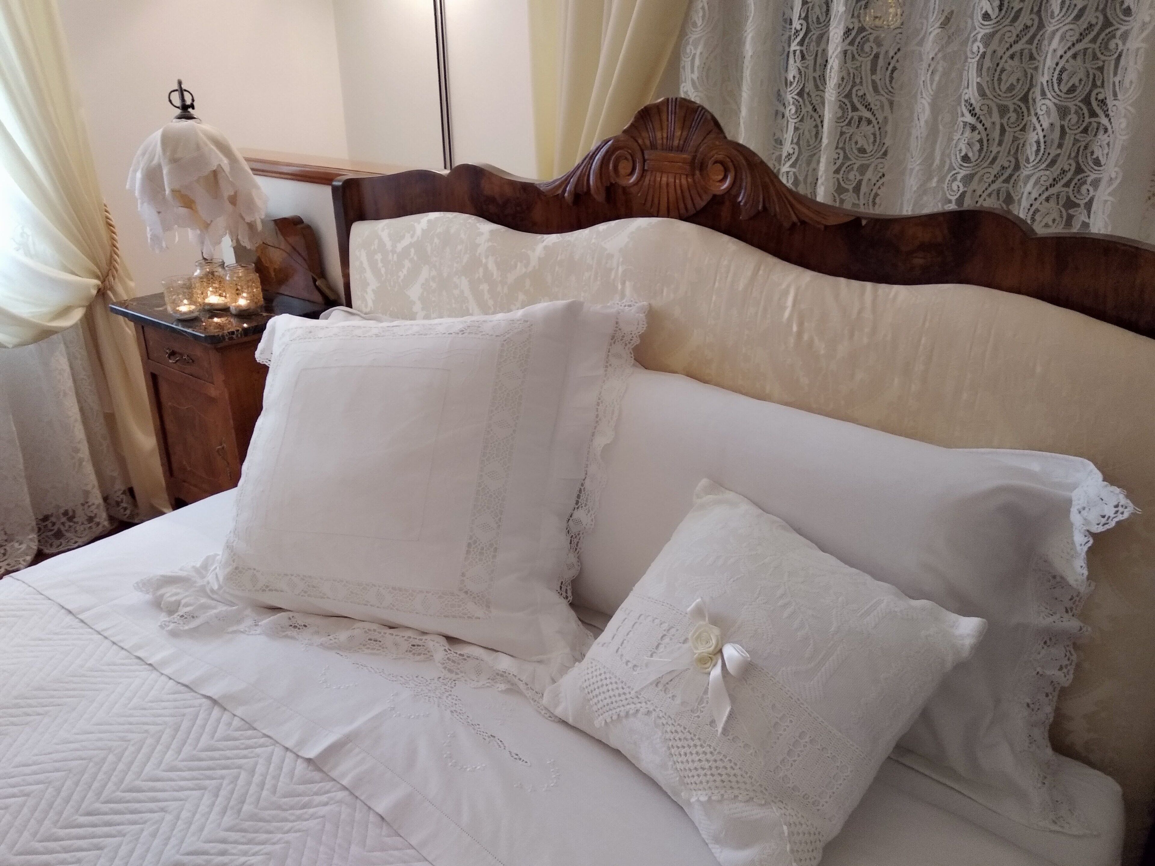 Una cama con sábanas blancas y detalles de encaje, un cabecero de madera y una mesita de noche con una lámpara y velas.