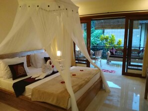 A bedroom with a four-poster bed, a balcony with outdoor furniture, and a view of greenery. - Qiul Guest House (Ubud)