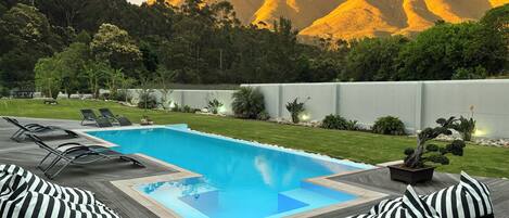 Una piscina con sillones de playa y cojines a rayas, rodeada de un césped cuidado y con montañas al fondo.