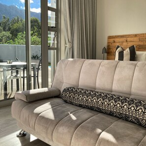 A modern living room with a beige sofa, a patterned throw pillow, and a view of mountains through large windows. - A Riverbed Guesthouse (Swellendam)