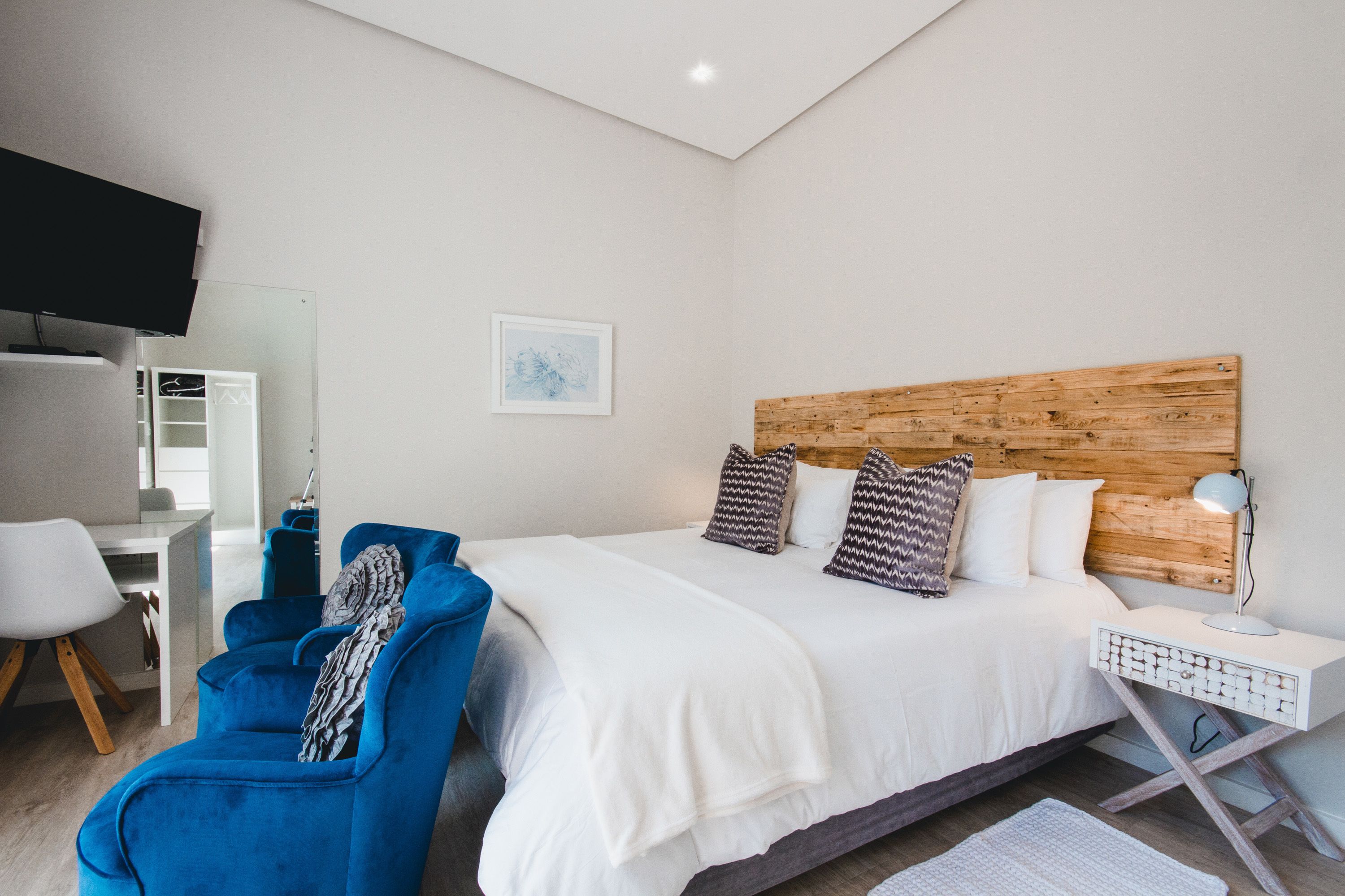 A modern bedroom with a wooden headboard, a large bed with white bedding, a blue armchair, a desk with a chair, and a flat-screen TV mounted on the wall.