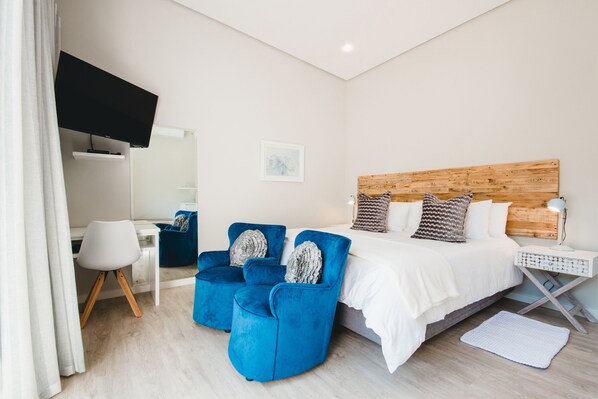 A modern hotel room with a bed, two blue armchairs, a desk, and a TV mounted on the wall. - A Riverbed Guesthouse (Swellendam)
