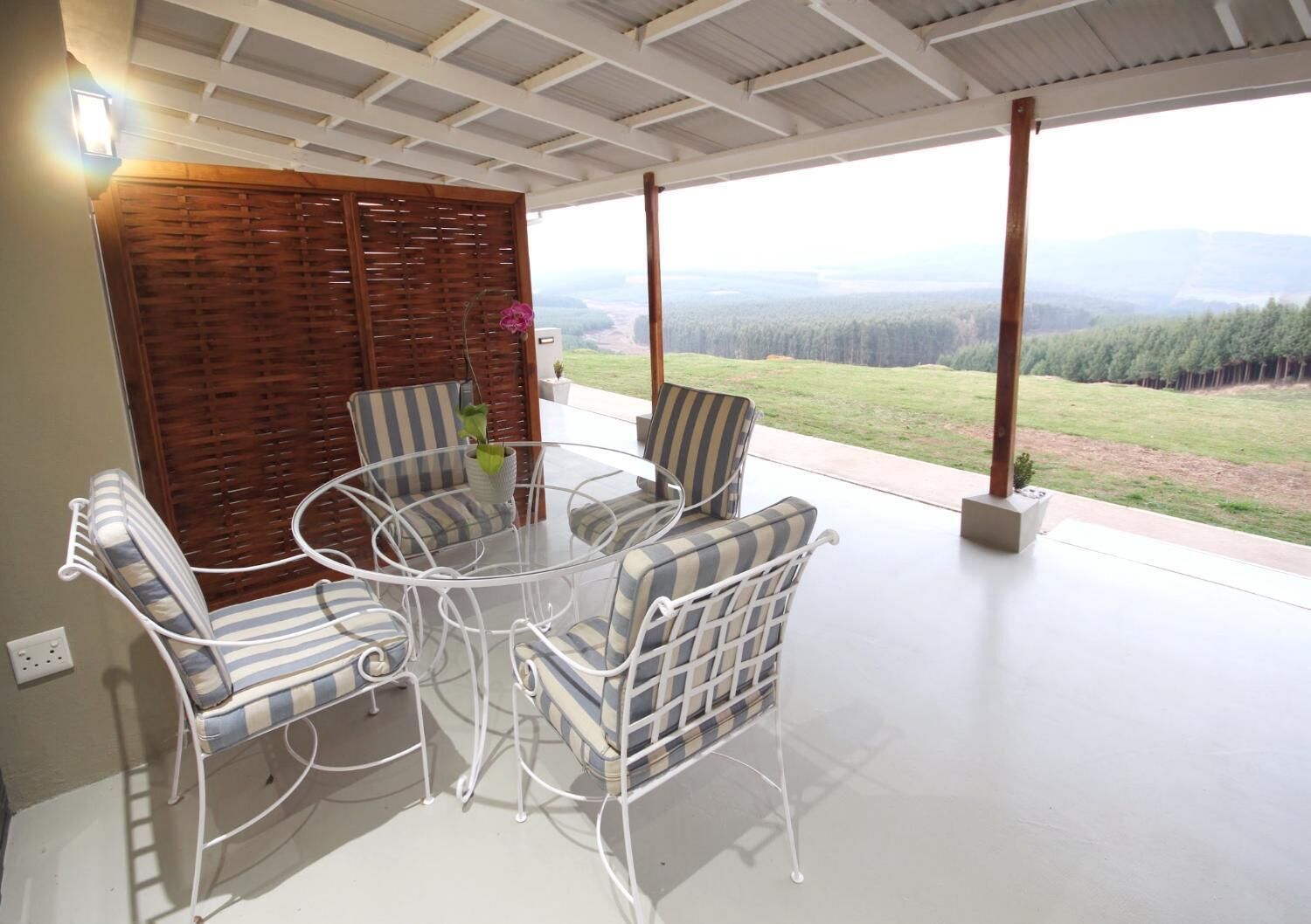 Un espace extérieur abrité, aménagé avec une table et des chaises, une cloison en bois et offrant une vue sur un paysage boisé.