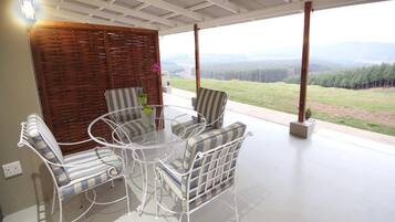 Un espace extérieur abrité, aménagé avec une table et des chaises, une cloison en bois et offrant une vue sur un paysage boisé.