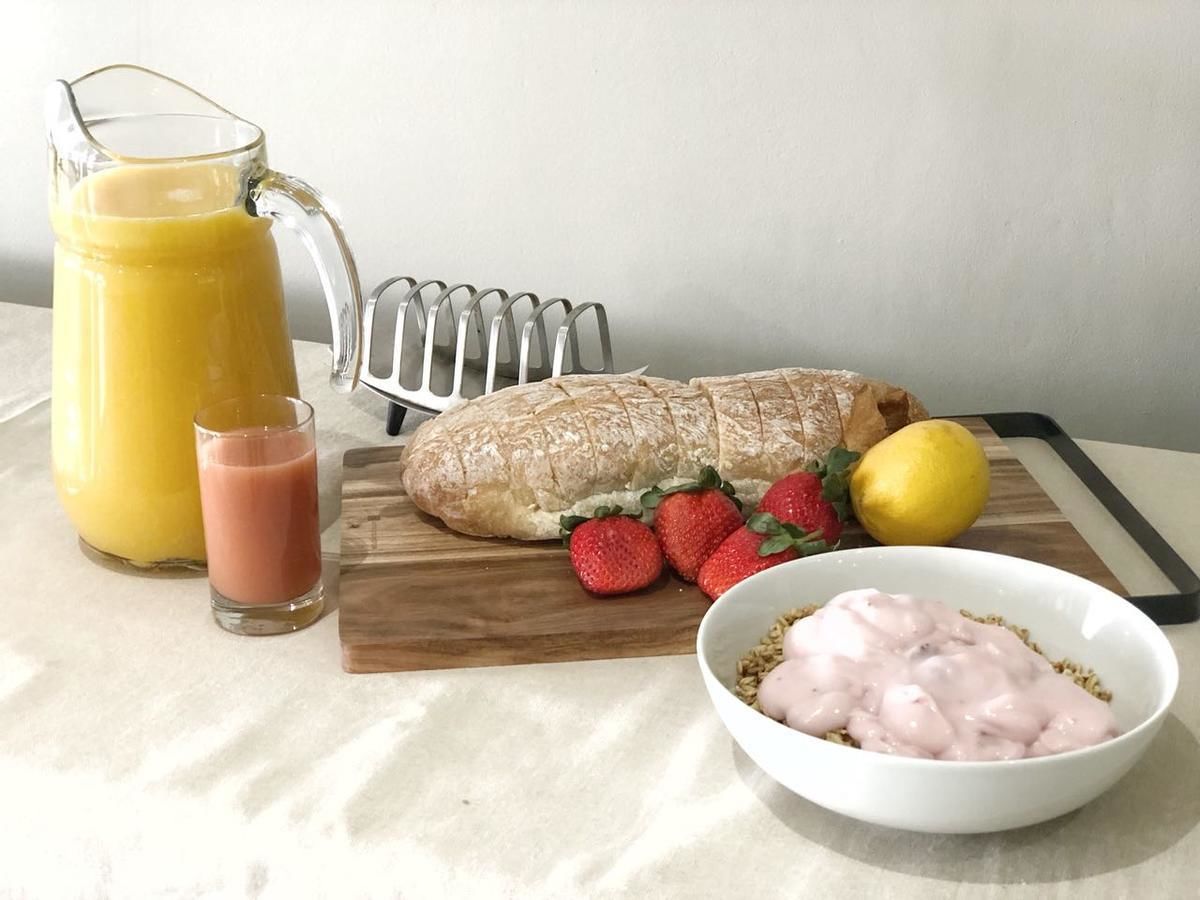 A breakfast setting with a baguette, strawberries, a glass of orange juice, and a bowl of cereal with yogurt.