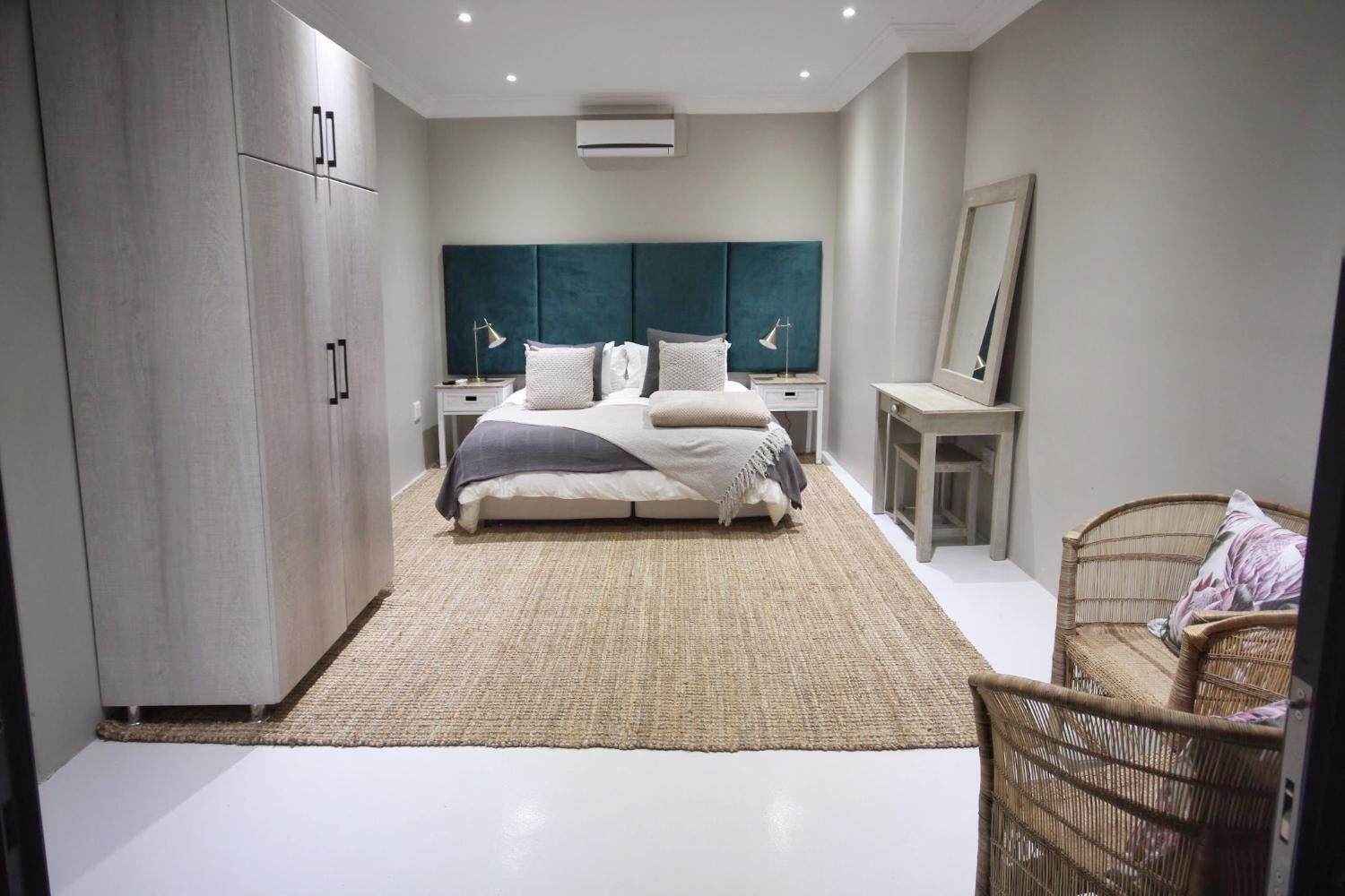 A modern bedroom with a large bed, a wardrobe, a bedside table, a mirror, and a wicker chair.