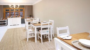 A dining area with a wooden table and white chairs, a kitchen area with a white cabinet, and a large window with curtains. - Curry's Post Bed and Breakfast (Curry's Post)