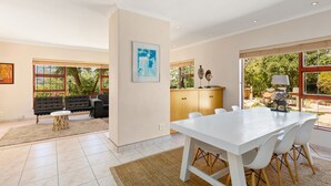 A spacious living area with a white dining table, black sofa, and a large window offering a view of greenery. - Hout Bay Hilltop - In Cape Town (Hout Bay) (Cape Town)